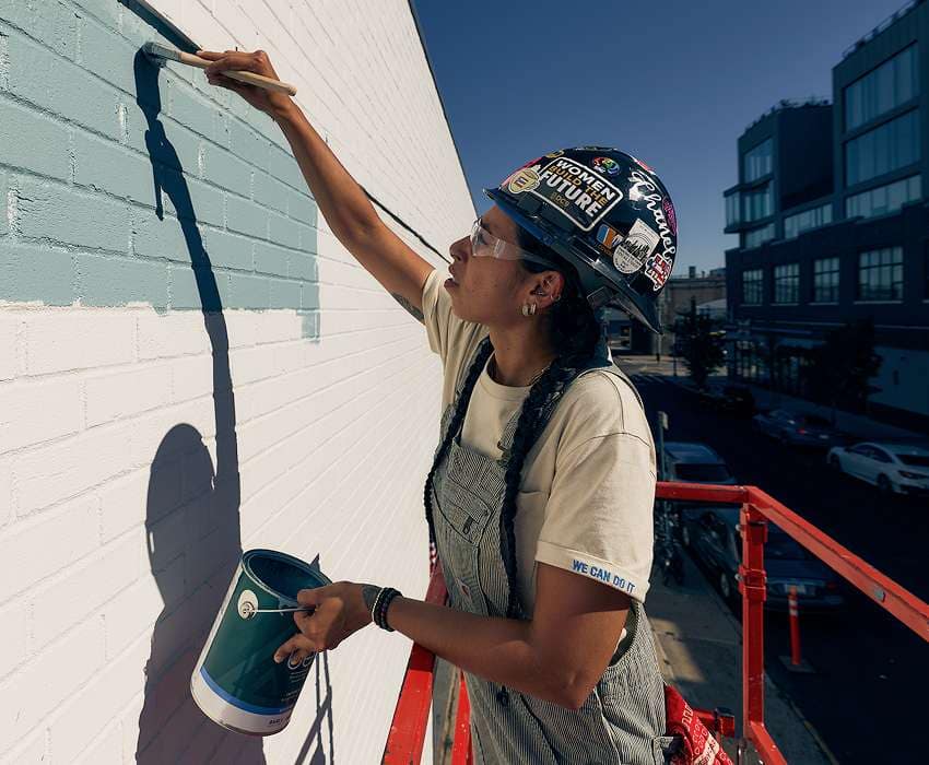 Female painter wearing a Carhartt tee and overalls painting the side of a building.