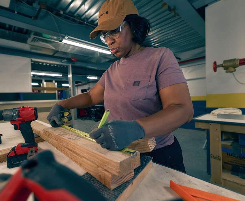 A female carpenter in safety glasses and a Carhartt tee and hat measuring lumber.