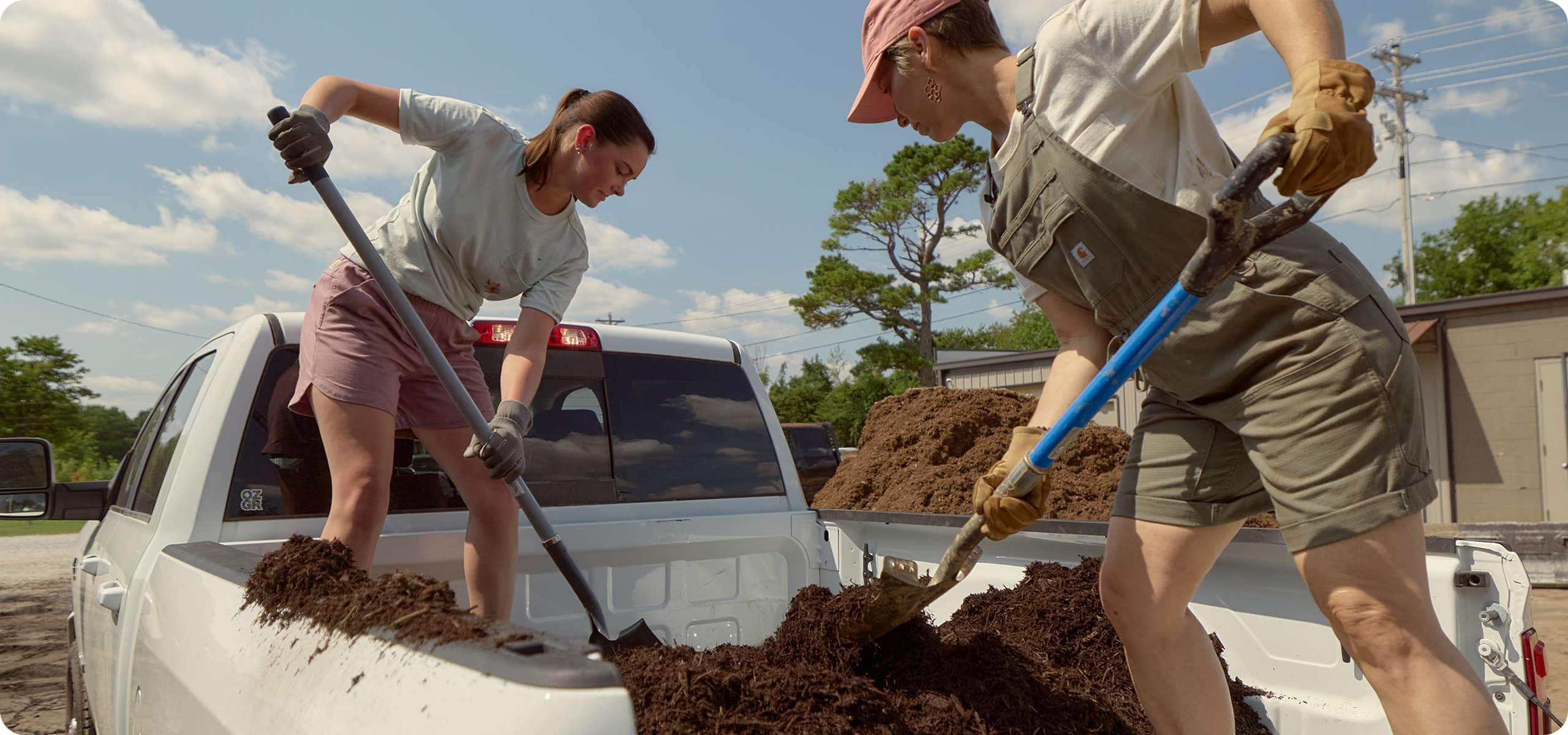 Two women wearing Carhartt warm weather gardening gear while shoveling soil from the back of a pickup truck.