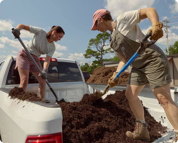 Two women wearing Carhartt warm weather gardening gear while shoveling soil from the back of a pickup truck.