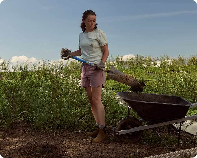 Emily of Ozark Green Roofs shoveling soil into a wheelbarrow while wearing a Carhartt tee, shorts, and gloves.