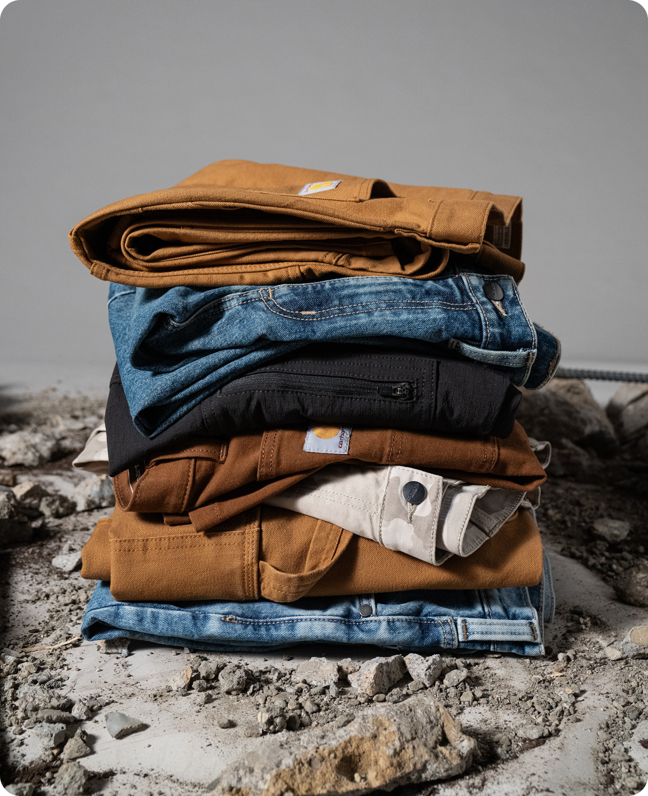 Carhartt work pants in different colors, styles, and materials folded and stacked on a rough, rocky surface