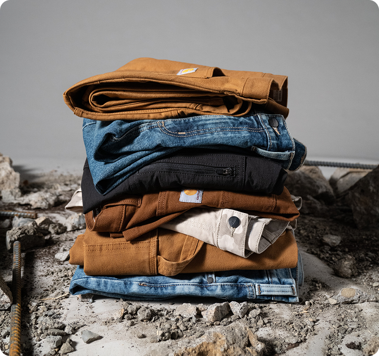 Carhartt work pants in different colors, styles, and materials folded and stacked on a rough, rocky surface